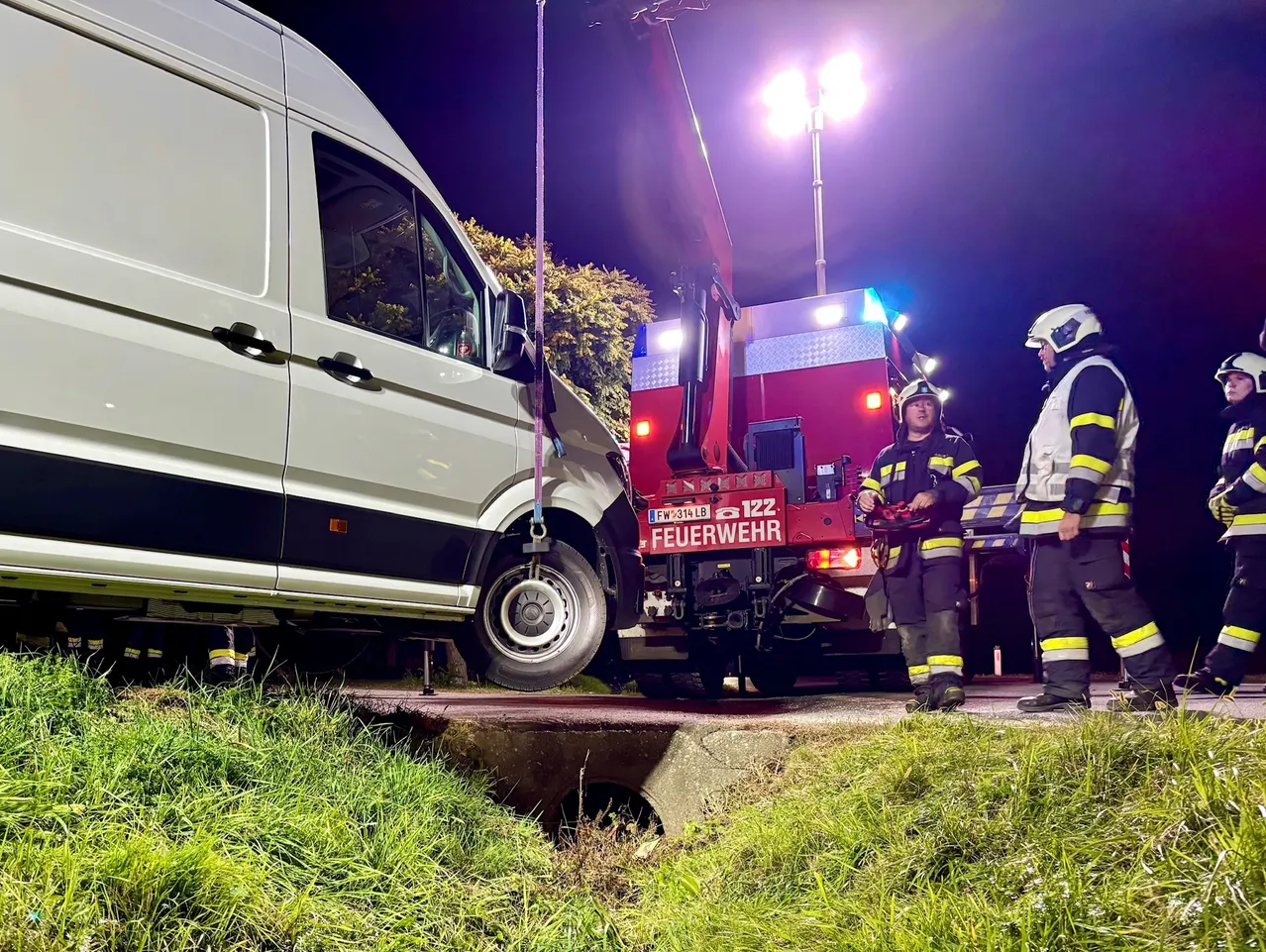 Ein weißer Lieferwagen steckt in einem Loch am Straßenrand. Ein Feuerwehrwagen steht dahinter mit eingeschalteten Lichtern. Vier Feuerwehrleute stehen um den Wagen herum, zwei von ihnen tragen Helme.