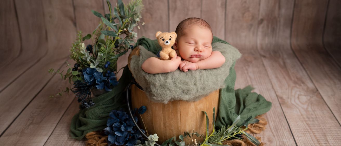 Ein Neugeborenes schläft in einem Holzeimer, umgeben von blauen und grünen Pflanzen. Ein brauner Teddybär liegt neben dem Baby. Die Umgebung hat einen rustikalen, erdigen Ton.