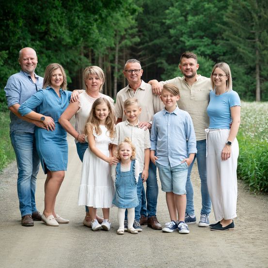 Eine Familie mit acht Personen steht lächelnd für ein Foto auf einem Kiesweg in einem bewaldeten Gebiet. Sie sind lässig gekleidet und der Hintergrund ist mit Bäumen und Blumen gefüllt.