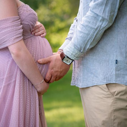 Eine schwangere Frau in einem lila Kleid hält Händchen mit einem Mann in einem blauen Hemd und beigen Hosen, die auf einem grünen Rasen stehen.