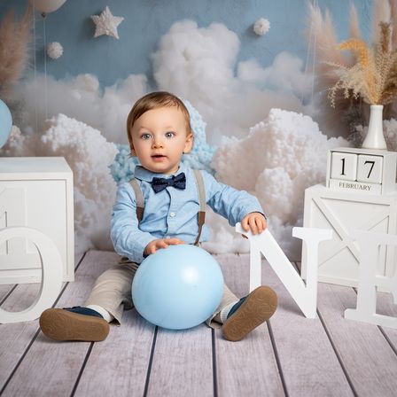 Ein Babyjunge sitzt auf einem Holzboden und trägt ein blaues Hemd und Hosenträger. Er hält einen blauen Ballon und schaut nach vorne. Der Hintergrund zeigt weiße Wolken und die Zahl 17 auf einem Kalender.