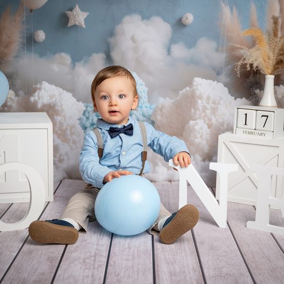Ein Babyjunge sitzt auf einem Holzboden und trägt ein blaues Hemd und Hosenträger. Er hält einen blauen Ballon und schaut nach vorne. Der Hintergrund zeigt weiße Wolken und die Zahl 17 auf einem Kalender.