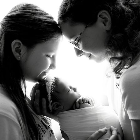Ein schwarz-weißes Foto von zwei Frauen und einem Neugeborenen. Das Baby ist in weißem Stoff gewickelt und wird von einer der Frauen gehalten.
