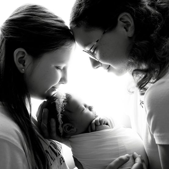 Ein schwarz-weißes Foto von zwei Frauen und einem Neugeborenen. Das Baby ist in weißem Stoff gewickelt und wird von einer der Frauen gehalten.