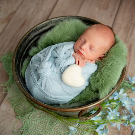 Ein Neugeborenes schläft in einem Eimer mit grünem Fell und einer blauen Decke. Das Baby hält ein weißes Objekt. Der Eimer steht auf einer Holzoberfläche.