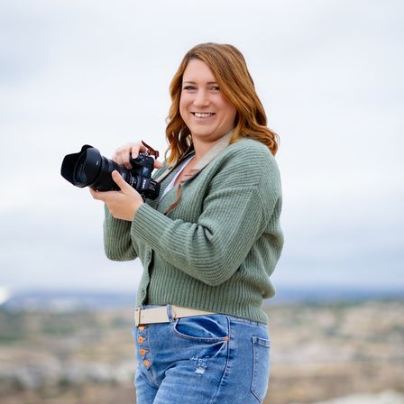 Eine Frau steht im Freien, lächelt und hält eine Kamera mit einer Umhängetasche um den Hals. Sie trägt einen grünen Cardigan und blaue Jeans. Der Hintergrund zeigt eine hügelige Landschaft.