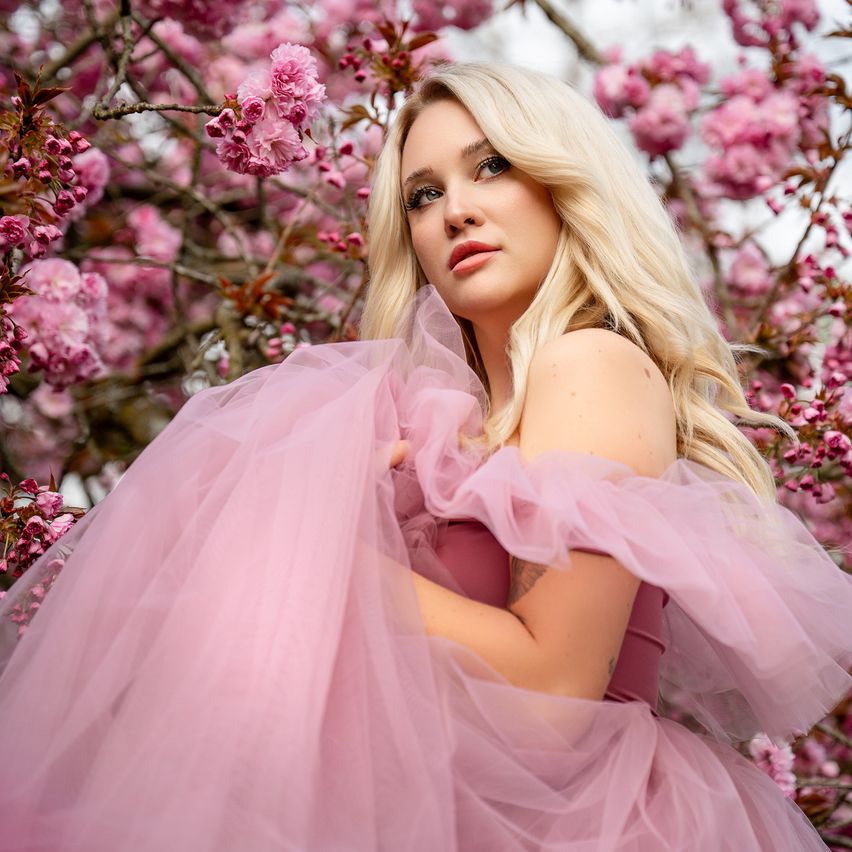 A woman with blonde hair poses under a cherry blossom tree, wearing a pink tulle dress. The background is filled with pink flowers, and she has a tattoo on her arm.