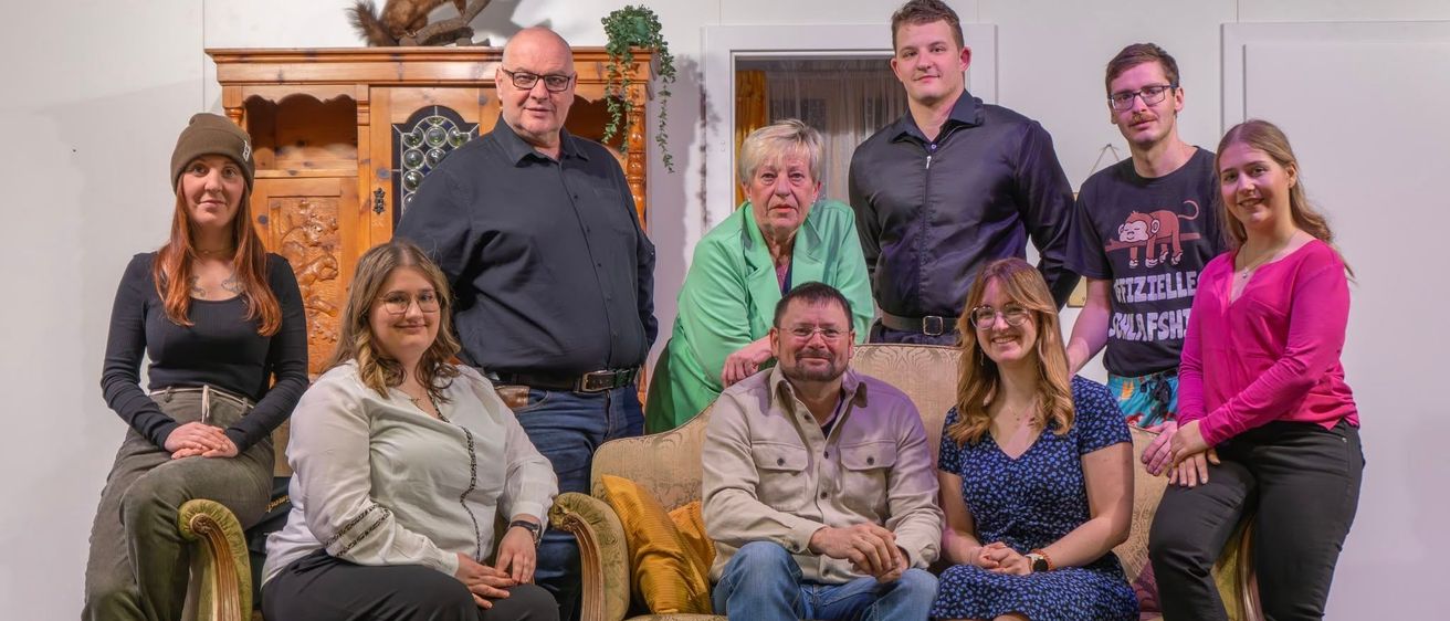 Eine Gruppe von Menschen sitzt auf einer Couch, einige stehen dahinter, in einem Raum mit einem Holzschrank, einer Pflanze und einer offenen Tür. Sie lächeln für ein Foto.