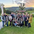 Eine Gruppe von Menschen posiert für ein Foto auf einem grünen Feld mit Bergen im Hintergrund. Einige sitzen, andere stehen hinter einem Baum.