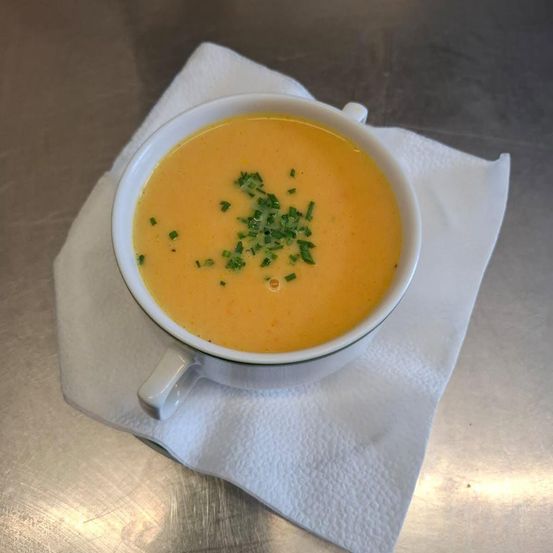 Eine Schüssel mit cremiger orangefarbener Suppe, garniert mit grünen Kräutern, steht auf einem weißen Papiertuch auf einer Metalloberfläche.