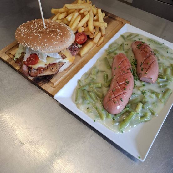 Ein Holzbrett hält einen Burger mit Pommes und ein weißer Teller mit zwei Würsten und grünen Bohnen.