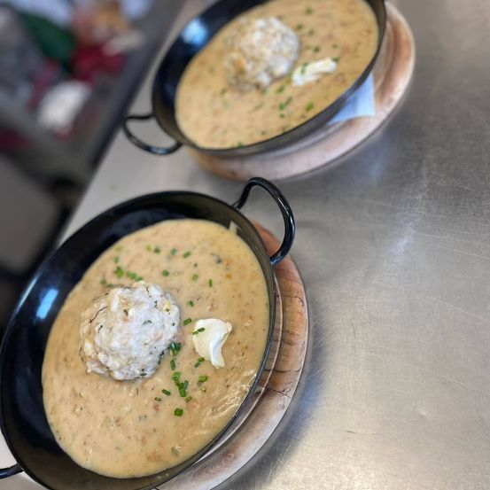 Zwei Schüsseln mit cremiger Suppe mit einer Fleischkugel und einem Butterklecks auf einem Holzteller auf einer Metalloberfläche.