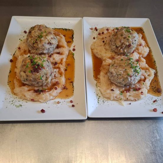 Zwei Teller mit Fleischbällchen auf Kartoffelpüree mit Soße, garniert mit Kräutern und Speckstückchen.