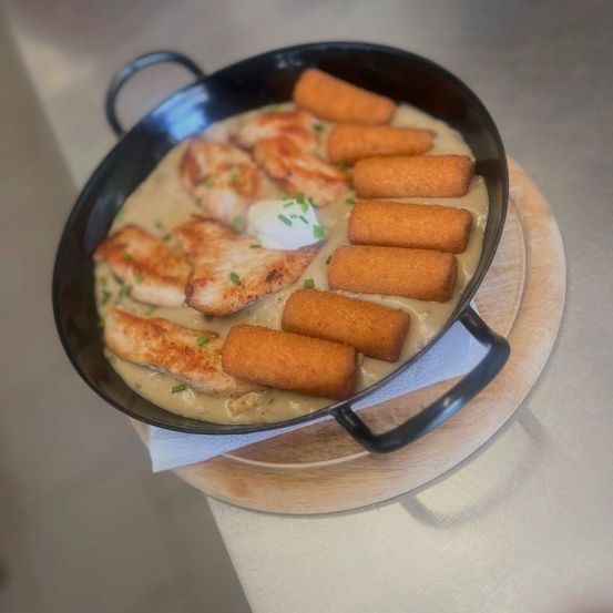 Eine schwarze Pfanne mit einem cremigen Gericht, das Hähnchen und mit Käse gefüllte Brötchen enthält, garniert mit grünen Kräutern, auf einem Holzteller.