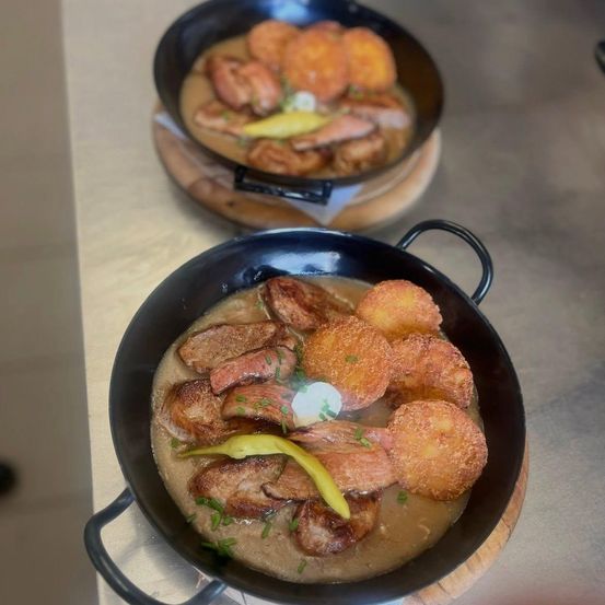 Zwei Pfannen mit einem Fleischgericht in Soße und frittierten Kartoffelbällchen, garniert mit grünen Kräutern und einer Zitronenscheibe.