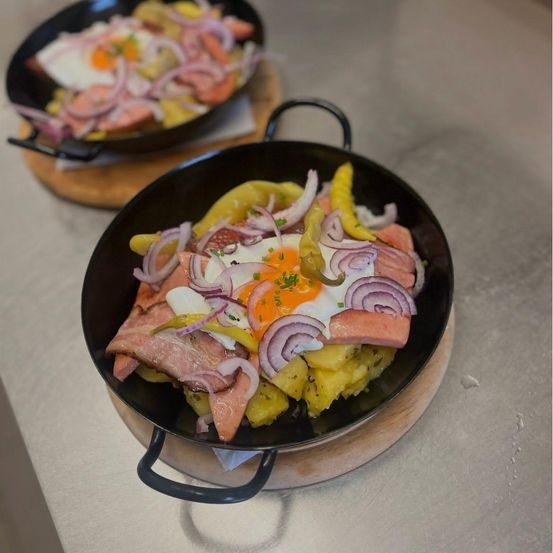Zwei schwarze Bratpfannen mit einer herzhaften Mahlzeit aus Kartoffeln, Zwiebeln und Schinken, gekrönt von einem Spiegelei, serviert auf Holzbrettern auf einer Metalloberfläche.