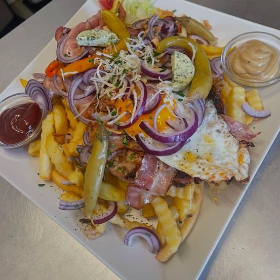 Ein Teller mit einem gebratenen Ei, Speck und Pommes, überzogen mit Zwiebeln, Paprika und Käse. Beilagen sind Ketchup und Senf.