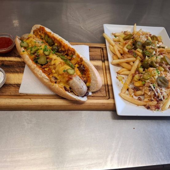 Ein Holztablett hält einen Hot Dog mit Käse, Gurken und Frühlingszwiebeln und eine Platte mit Pommes mit Speck, Käse und Gurken.