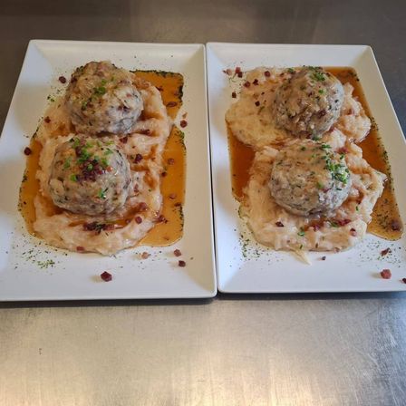 Zwei Teller mit Fleischbällchen in cremiger Sauce mit Kartoffelpüree, garniert mit Kräutern und Speckstückchen.