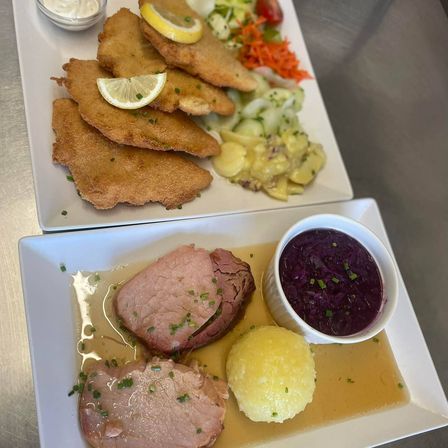 Zwei weiße Teller mit traditionellen deutschen Gerichten. Der obere Teller hat gebratenes paniertes Fleisch mit einer Zitronenscheibe und gemischtem Gemüse. Der untere Teller hat eine Scheibe Schinken, Kartoffelknödel und eine Schüssel Rotkohl.