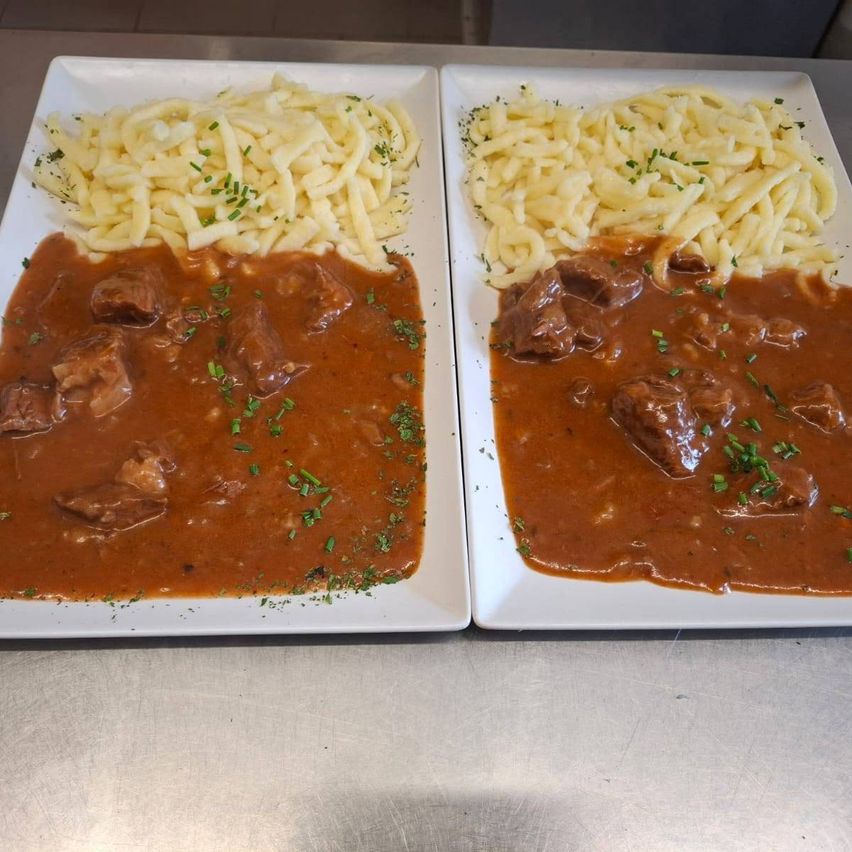 Two plates of beef stew with gravy and noodles garnished with green herbs on a metal surface.