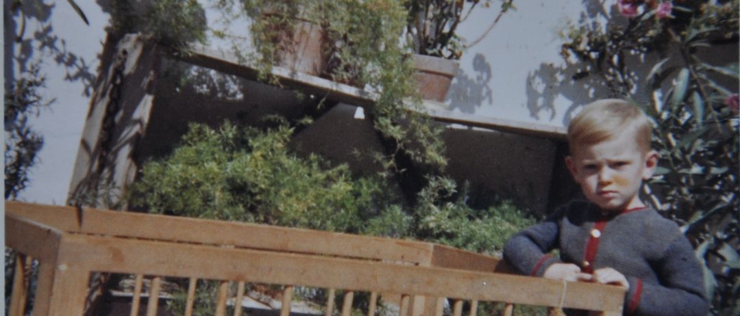 Ein altes Foto zeigt einen kleinen Jungen, der vor einem hölzernen Kinderbett steht, in dem ein schlafendes Baby liegt. Hinter ihnen stehen Blumentöpfe mit rosa Blumen auf einem Regal.