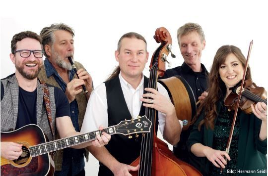 Sechs Musiker posieren für ein Foto, halten Gitarren, eine Geige und einen Cello. Sie alle lächeln und ein Mann bläst in eine Flöte.