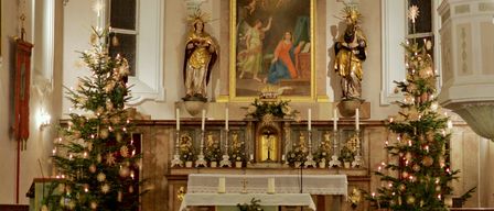 Ein geschmückter Kirchenaltar mit zwei Statuen, einem Gemälde und einem Weihnachtsbaum im Vordergrund. Der Altar hat Blumen, Kerzen und einen Tisch mit einer weißen Tischdecke.