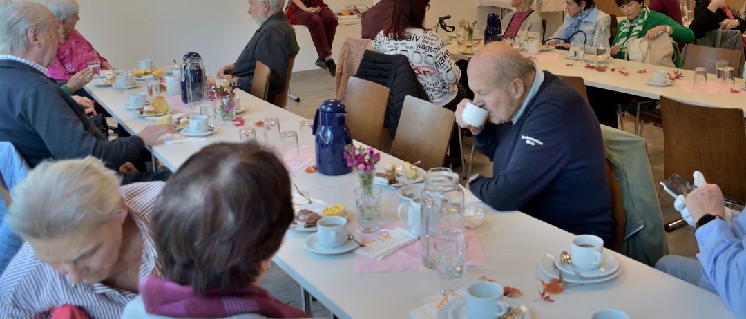 Eine Gruppe älterer Menschen sitzt um einen Tisch herum, einige trinken Kaffee. Der Tisch ist mit Tellern, Tassen, Blumen und einem Krug gedeckt. Ein Mann auf der rechten Seite trinkt aus einer Tasse. Im Hintergrund sitzt eine Frau auf einer Bank.