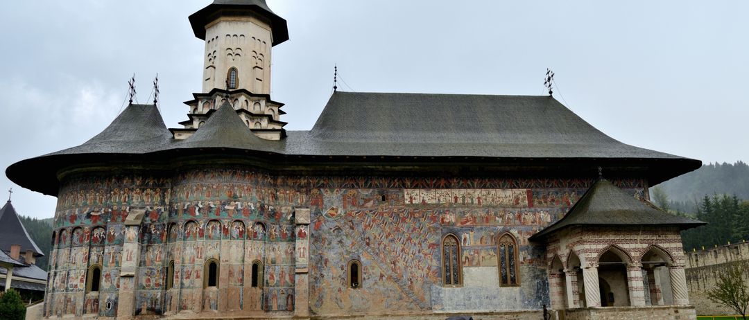 Ein Paar geht mit einem Schirm vor einer historischen Kirche mit einem Kirchturm und detaillierten Wandgemälden.