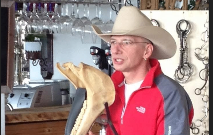 Ein Mann mit Cowboyhut hält ein Fossil in einer Bar. Er trägt einen roten Mantel mit Logo auf der linken Seite. Hinter ihm sind Weingläser und Metallketten.
