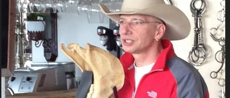 A man wearing a cowboy hat holds a fossil in a bar. He wears a red jacket with a logo on the left side. Behind him are wine glasses and metal chains.