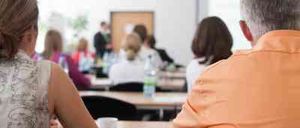 Eine Gruppe von Menschen sitzt in einem Klassenzimmer und schaut auf einen Redner vorne. Eine Frau in der ersten Reihe schaut zurück, während ein Mann neben ihr den Redner beobachtet. Flaschen und Papiere liegen auf dem Tisch.