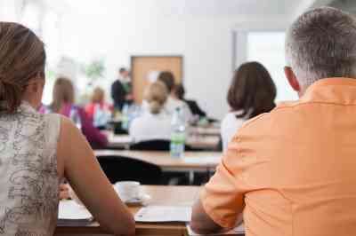 Eine Gruppe von Menschen sitzt in einem Klassenzimmer und schaut auf einen Redner vorne. Eine Frau in der ersten Reihe schaut zurück, während ein Mann neben ihr den Redner beobachtet. Flaschen und Papiere liegen auf dem Tisch.