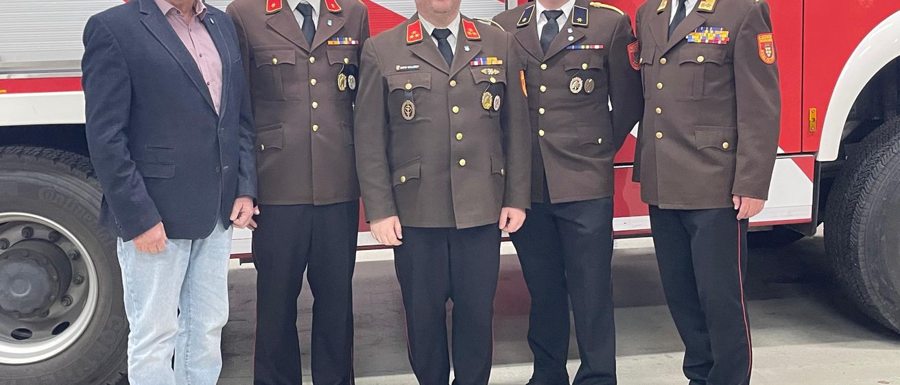 Fünf Männer stehen vor einem Feuerwehrauto. Sie tragen Uniformen und Lederschuhe. Drei von ihnen tragen Medaillen.