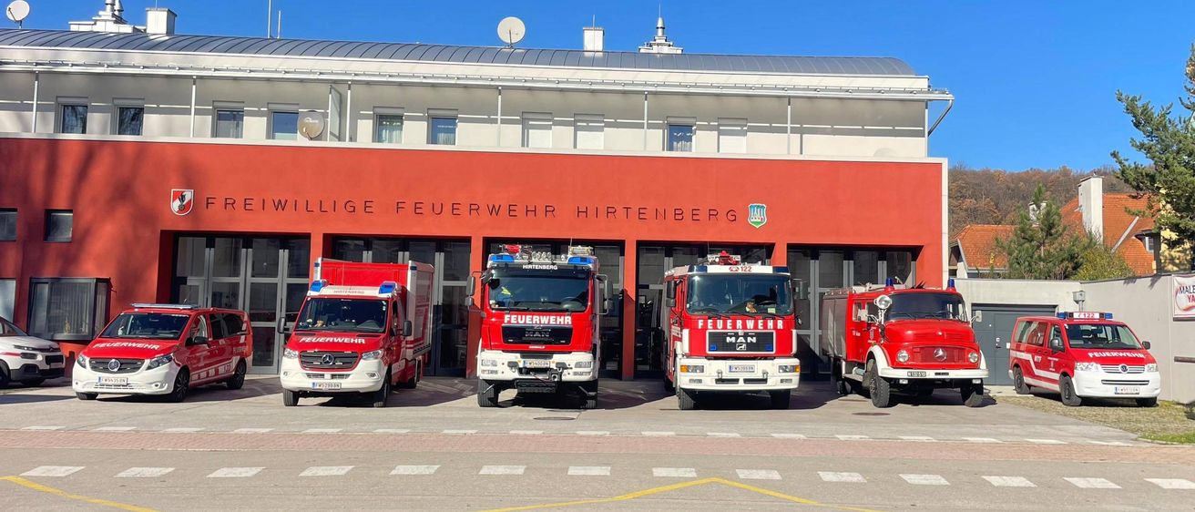 Drei rote Feuerwehrfahrzeuge sind vor dem Gebäude der Feuerwehr Hirtenberg geparkt. Das Gebäude hat eine rote Fassade mit weißen Fenstern und einem Logo.