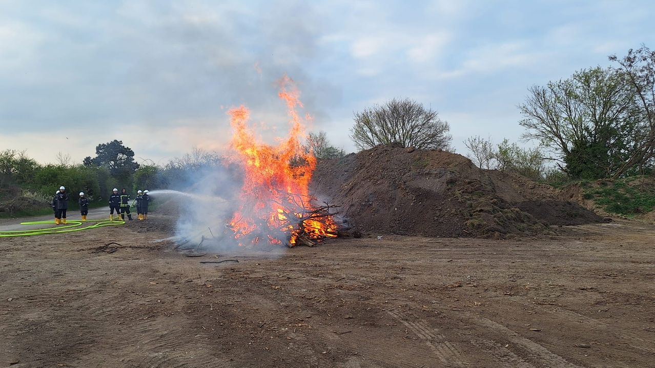 Ein Feuer brennt durch einen Haufen Holz in einem offenen Bereich, mit einem Feuerwehrmann, der in der Nähe einen Schlauch verwendet. Das Feuer ist intensiv und produziert viel Rauch.