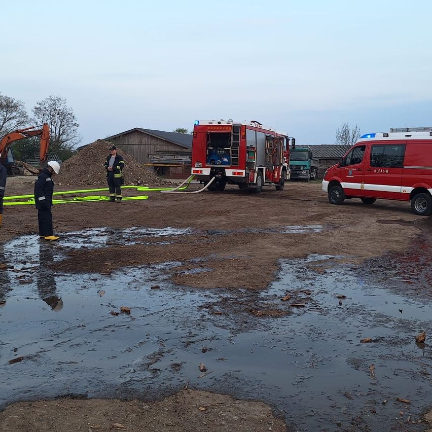 Feuerwehrleute arbeiten an einem Gelände mit Wasser und Ausrüstung. Ein roter Feuerwehrwagen ist geparkt und eine Person beobachtet.