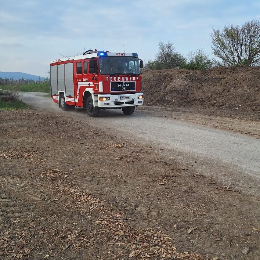 Ein roter Feuerwehrwagen mit blauer Licht auf der Oberseite fährt auf einer unbefestigten Straße. Der Wagen hat 'FEUERWEHR' und 'MAN' vorne geschrieben.