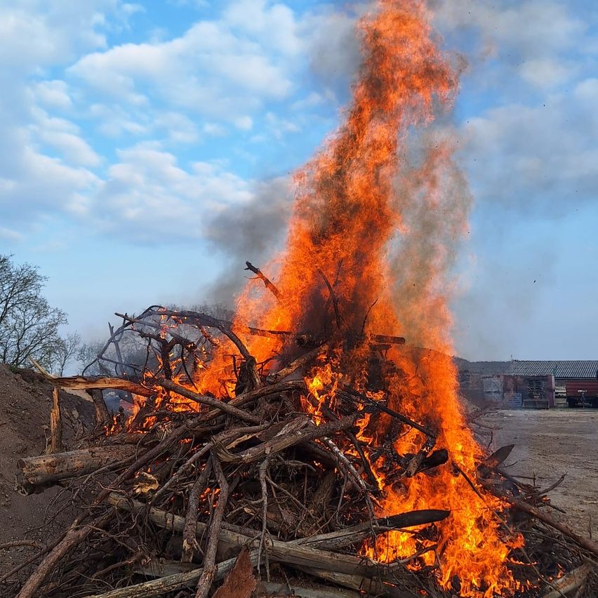 Ein großes Lagerfeuer brennt hell und ist von einem Haufen Holz und Zweigen umgeben. Das Feuer wirft einen warmen Schein auf den Boden, und der Himmel ist klar mit verstreuten Wolken.