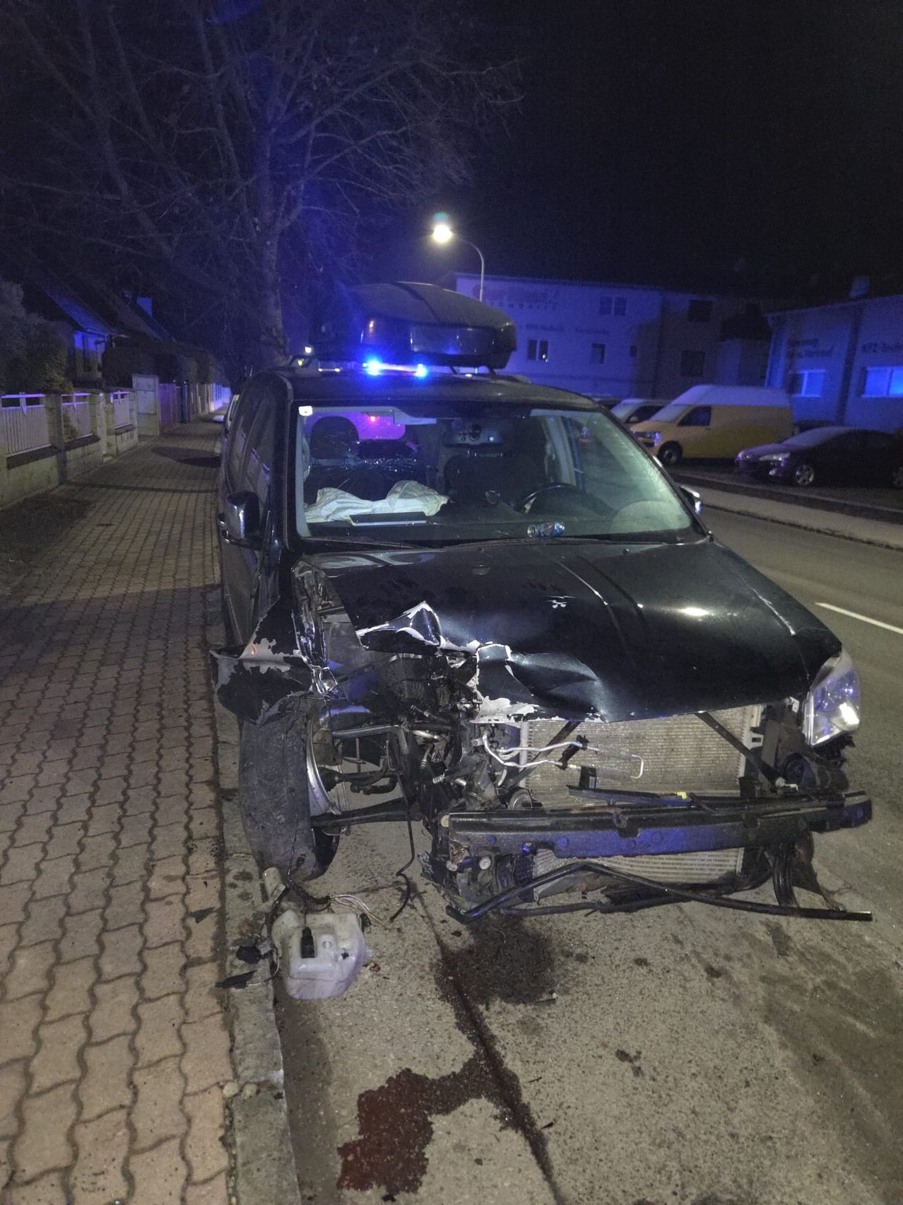 Ein schwarzer SUV mit eingeschalteten Blaulichtern steht nachts auf einem Bürgersteig, vorne beschädigt.