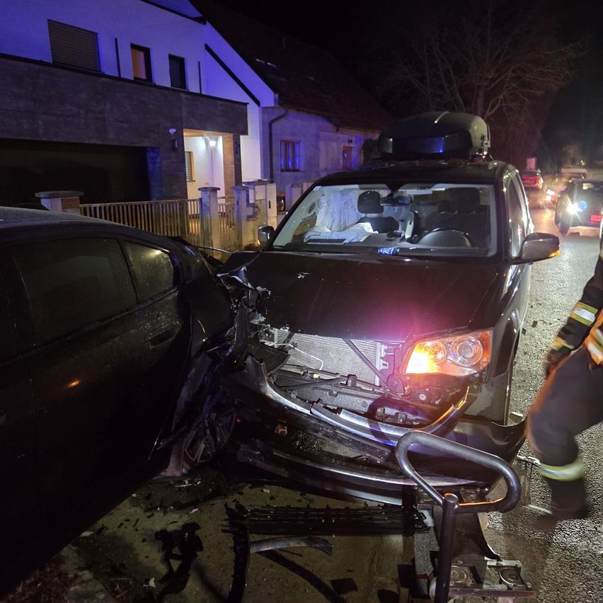 Ein nächtlicher Autounfall zeigt einen schwarzen SUV mit vorderer Beschädigung und einem Feuerwehrmann in der Nähe. Ein weiteres Auto ist ebenfalls beschädigt, und ein Haus mit beleuchteten Fenstern ist im Hintergrund.
