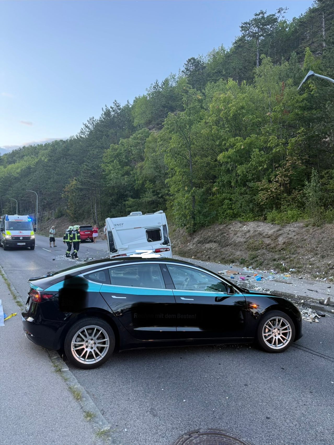 Ein Tesla Model 3 steht am Straßenrand, neben ihm sind Rettungsfahrzeuge und Personen. Die Straße ist von Grün und Bäumen umgeben.
