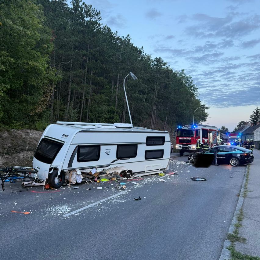 Ein weißer Wohnwagen ist auf der Straßenseite beschädigt, mit Rettungsfahrzeugen in der Nähe.