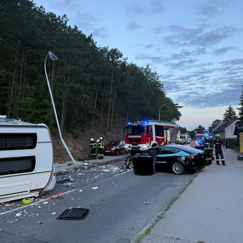 Ein Verkehrsunfall mit einem Tesla und einem Wohnwagen ereignete sich auf der Straße, Rettungskräfte sind vor Ort.