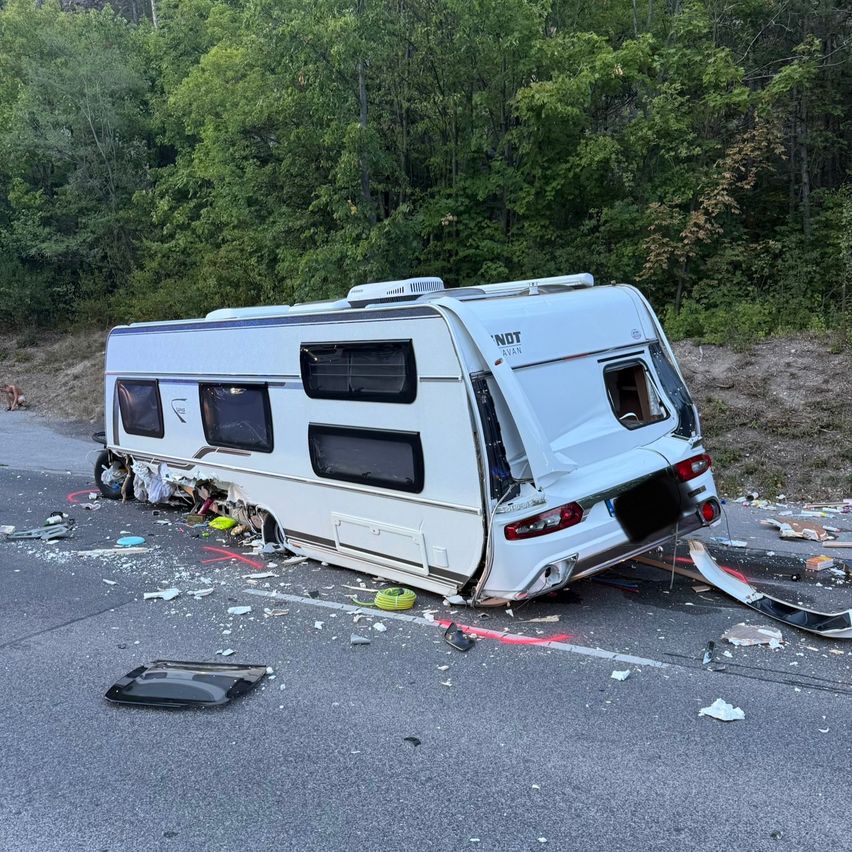 Ein weißer Wohnwagen ist auf der Straßenseite umgekippt, mit herumliegenden Trümmern. Bäume und ein Hügel sind im Hintergrund sichtbar.