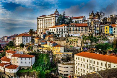 Ein Blick vom Hügel auf Porto, Portugal, zeigt eine lebendige Stadtlandschaft mit bunten Gebäuden, roten Dächern und üppigem Grün unter einem teilweise bewölkten Himmel.
