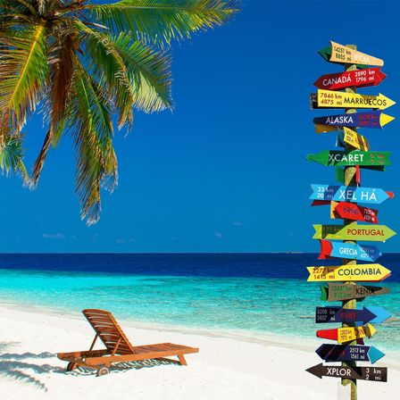 Ein hölzerner Wegweiser mit verschiedenen Schildern, die auf verschiedene Ziele wie Kanada, Indien, Alaska und Xcaret zeigen. Ein hölzerner Liegestuhl steht neben dem Wegweiser am Strand. Der Strand hat weißen Sand und klares blaues Wasser. Der Himmel ist klar und blau.