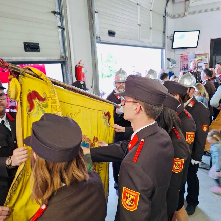 Eine Gruppe von Feuerwehrleuten ist in einer Feuerwache versammelt, ein Mann hält eine gelbe Fahne, während andere ihn betrachten.