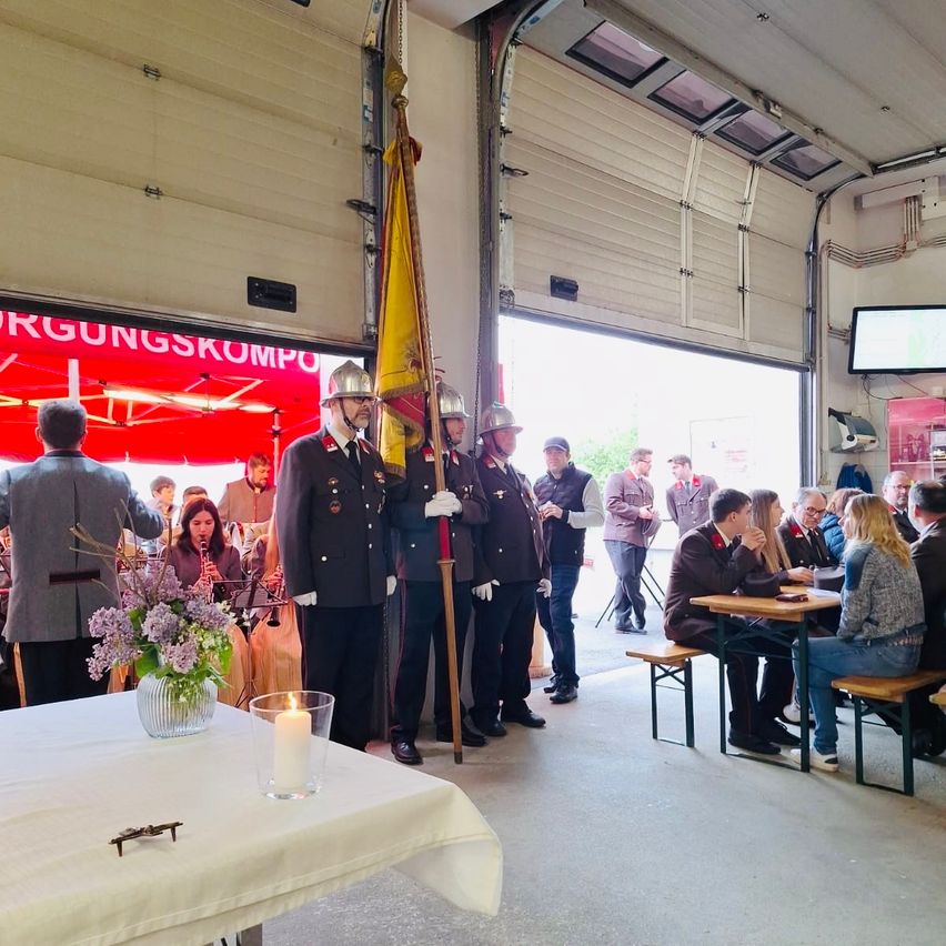 Mehrere uniformierte Feuerwehrleute stehen vor einem Gebäude mit einer Flagge, einer Kerze auf einem Tisch und Menschen, die an Tischen sitzen.